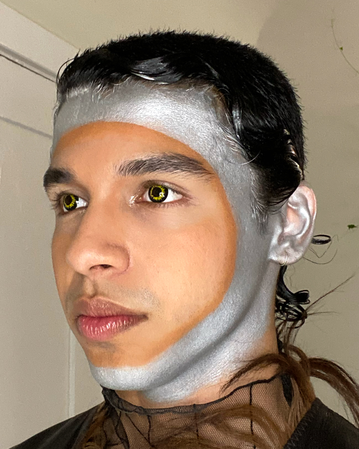 Model with silver makeup around the perimeter of his face against a tan background